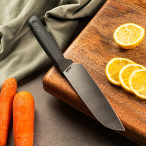 #15cm/6"
A Zyliss Santoku Knife with an ergonomic handle sits on a wooden cutting board beside three lemon slices, a halved lemon, two whole carrots, and a green cloth spread on the gray surface.