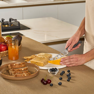 A person slices cheese with a Zyliss Cheese Knife 12.7cm/5", featuring a wheat straw handle and stainless steel blade, on a round white plate with crackers, beside bread, fruit, orange juice, blueberries, and cherries.