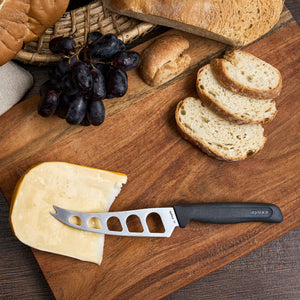 The Zyliss Cheese Knife 12.7cm/5" with a wheat straw handle and stainless steel blade rests on a wooden board alongside sliced bread, cheese, and grapes, while a basket filled with more bread is in the background.