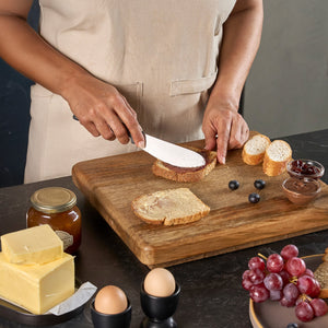A person uses the Zyliss Spreading Knife 12cm/5" to butter bread on a wooden cutting board, surrounded by sliced baguette, blueberries, jam, grapes, eggs, butter, and a jar of honey.