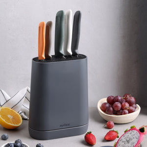 The Zyliss 6 Piece Universal Knife Block, featuring colorful, ergonomic handles, sits on a kitchen counter beside half an orange, a towel, a bowl of grapes, and sliced dragon fruit.