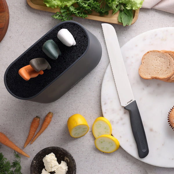 A Zyliss 6 Piece Universal Knife Block with four colorful stainless steel knives is displayed beside a serrated bread knife, an ergonomic handle, and a marble cutting board holding sliced bread, carrots, squash, and lettuce.