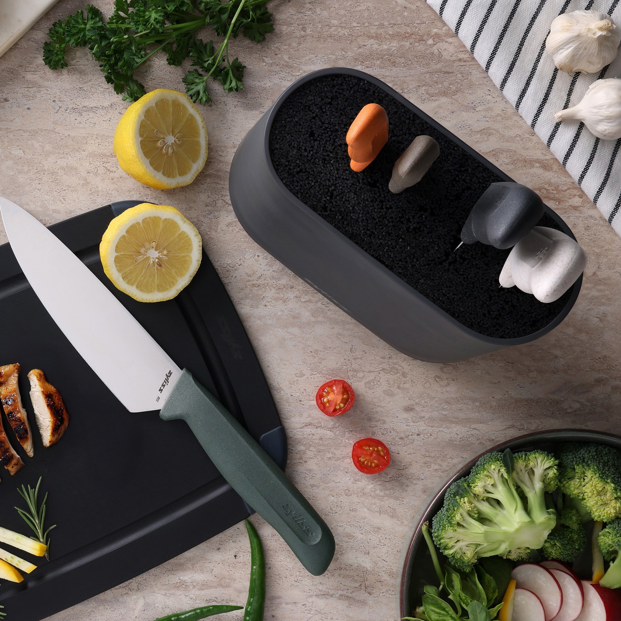On the kitchen counter, a Zyliss 6 Piece Universal Knife Block with stainless steel knives and ergonomic handles sits beside sliced lemon, grilled chicken, cherry tomatoes, broccoli, herbs, a striped towel, and garlic cloves.