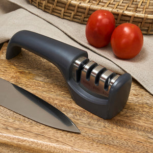 The Zyliss Knife Sharpener, featuring three slots for serrated edge care, sits on a wooden cutting board beside a kitchen knife, with two tomatoes and a woven basket in the background.