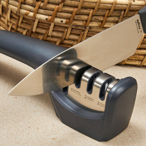 A kitchen knife is being sharpened with the Zyliss Knife Sharpener—a black and silver three-stage tool for kitchen blades and serrated edges—placed on a countertop with a woven basket in the background.