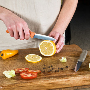 A person uses the Zyliss 2 Piece Knife Set to slice a lemon on a wooden cutting board; nearby are tomato slices, yellow pepper, lettuce, black peppercorns, and the other knife from the set with its blade guard.