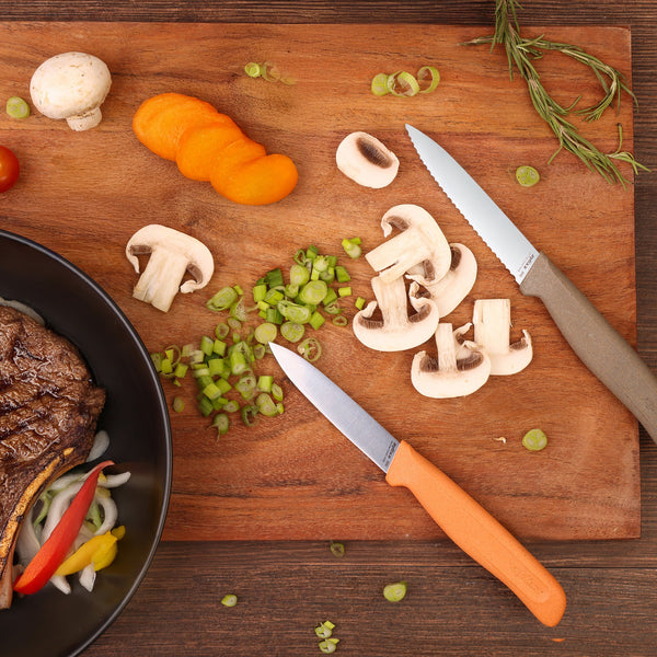 A Zyliss 2 Piece Knife Set rests on a wooden cutting board with sliced mushrooms, green onions, and an orange vegetable. Nearby, a black bowl holds cooked steak with onions and peppers. Rosemary and a whole mushroom are also visible.