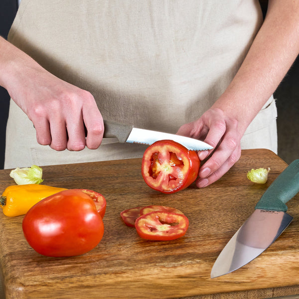 Wearing a beige apron, a person uses a green-handled knife from the Zyliss 3 Piece Knife Set to slice a tomato on a wooden cutting board, with a whole tomato and yellow pepper nearby.