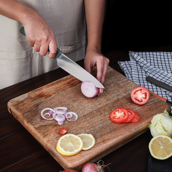 A person uses a Zyliss 3 Piece Knife Set to slice a red onion on a wooden cutting board, surrounded by sliced tomatoes, lemon, lettuce, and a checkered towelâ€”showcasing an organized kitchen setup.