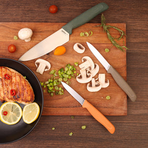 The Zyliss 3 Piece Knife Set is displayed on a wooden cutting board with chopped mushrooms, green onions, cherry tomatoes, rosemary, and grilled chicken garnished with lemon and chili slices on a dark wood surface.