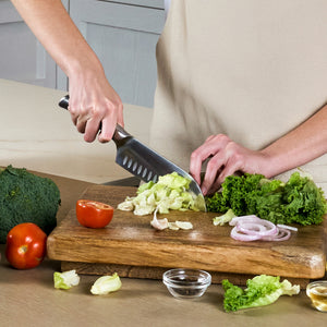 A person chops lettuce with the Zyliss Comfort Pro Santoku Knife 18.5cm/7", featuring an ergonomic handle, on a wooden cutting board amid tomato, broccoli, onion slices, and garlic in a kitchen setting.