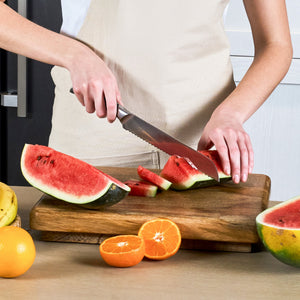 Wearing a beige apron, a person slices a watermelon on a wooden cutting board using the Zyliss Comfort Pro Bread Knife 21.5cm / 8½" with its serrated blade. Orange slices and assorted fruits are nearby.