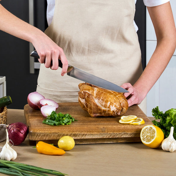 Wearing a beige apron, a person slices roast using the Zyliss Comfort Pro Carving Knife 20cm/8" with an ergonomic handle on a wooden board, surrounded by fresh veggies, greens, lemon halves, onions, and garlic on the kitchen counter.