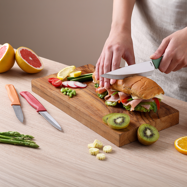 A person slices a deli meat and veggie sandwich on a wooden cutting board amid kiwi, grapefruit, asparagus, and lemon. Nearby rests the Zyliss Classic 3-Piece Paring & Utility Knife Set, ready for precise food prep.