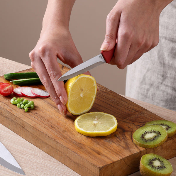 Using the Zyliss Classic 3-Piece Paring & Utility Knife Set, a person slices a lemon on a wooden cutting board surrounded by cut kiwi, cucumber, radish, tomato, and green beans.