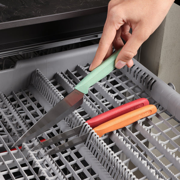 A hand places a knife from the Zyliss Classic 3-Piece Paring & Utility Knife Set with a green handle blade-down into a dishwasher rack, beside two other stainless steel knives with orange and red handles.