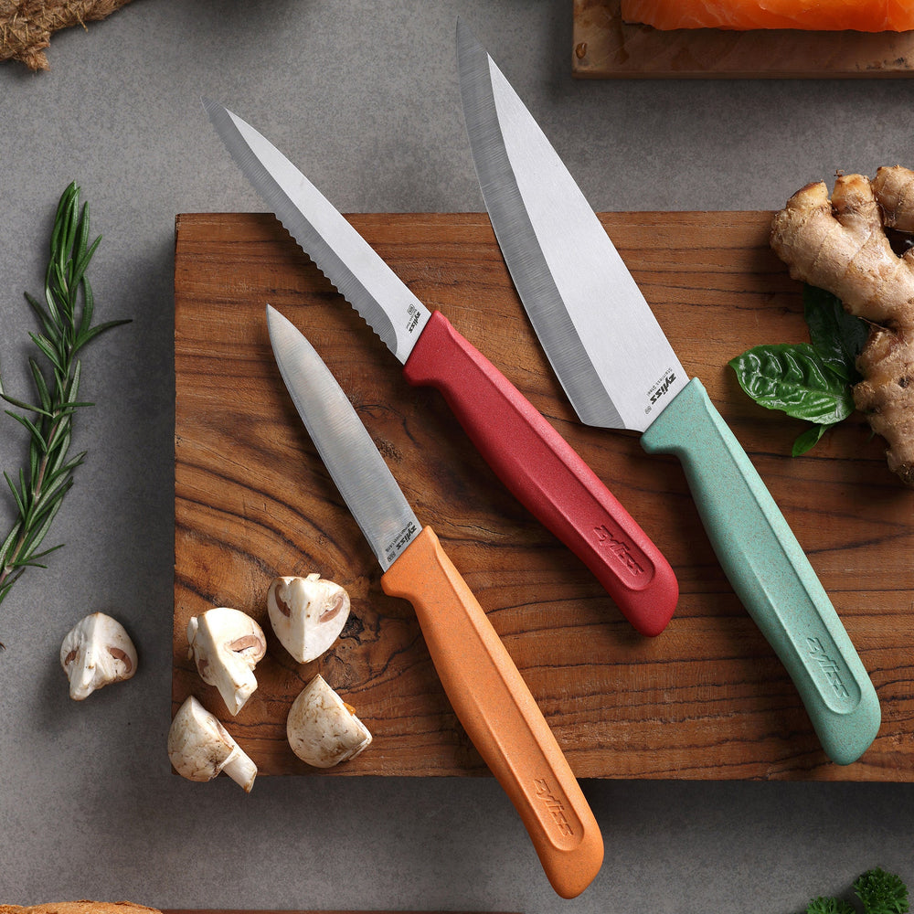The Zyliss Classic 3-Piece Paring & Utility Knife Set features stainless steel knives with colorful orange, red, and green handles. Upright against a light background, the center utility knife is serrated; the paring knives have smooth blades.