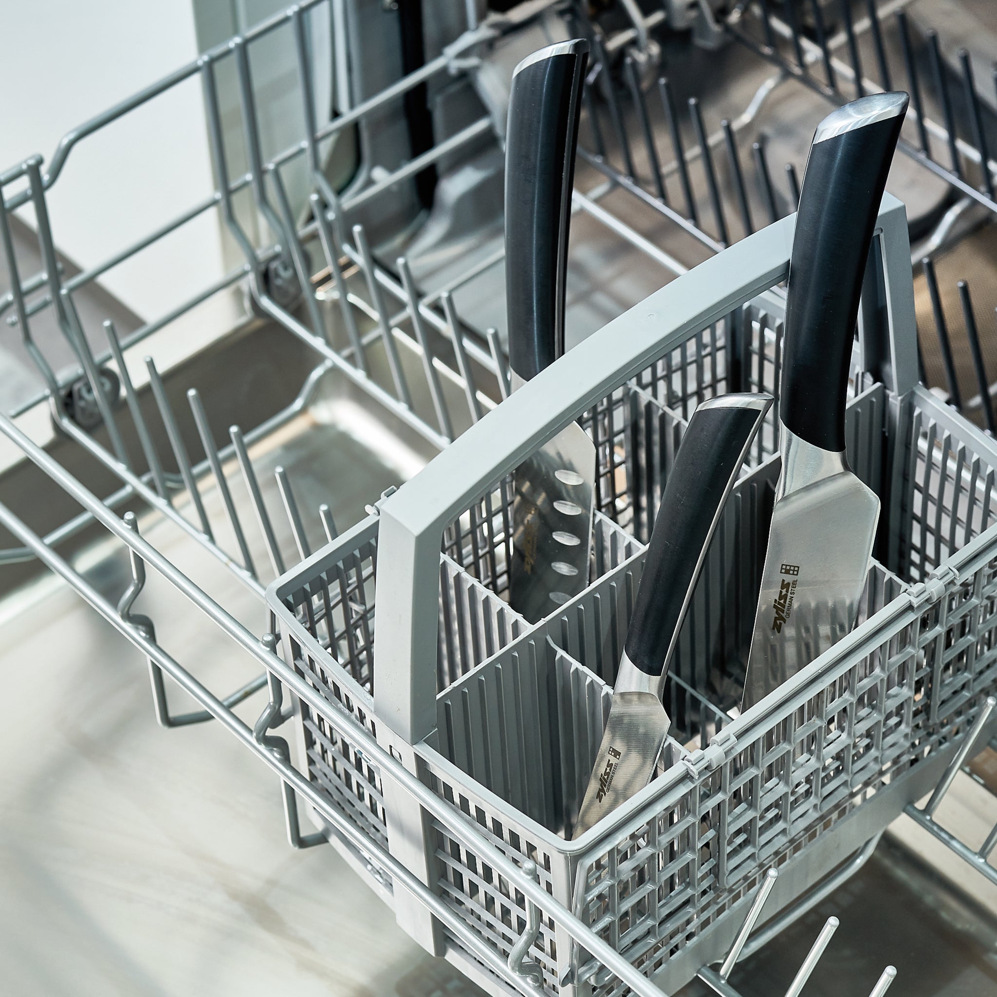 Three large kitchen knives are placed blade-up in a dishwashers cutlery basket, inside the lower rack. The dishwasher is open and empty apart from the knives.