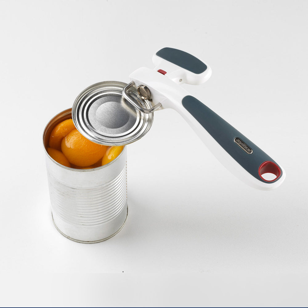 The Zyliss Safe Edge Can Opener has an ergonomic design, white and dark gray handle with red accents, and a metal turning mechanism, displayed against a plain light background. 