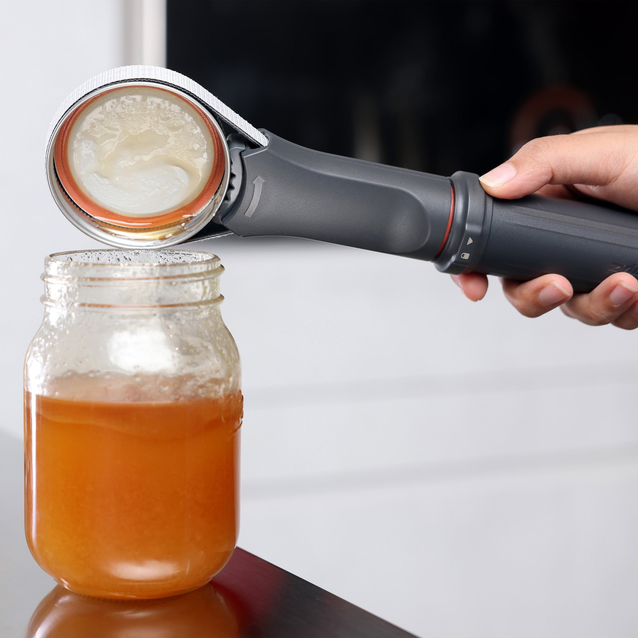A hand uses the Zyliss Jar Opener to grip and lift the lid off a glass jar filled with brown liquid.