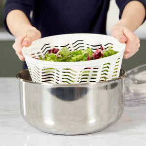 Someone lifts a white plastic basket of greens from the Zyliss Easy Spin 2 Stainless Steel Salad Spinner, which features a non-slip base, on a kitchen counter. 