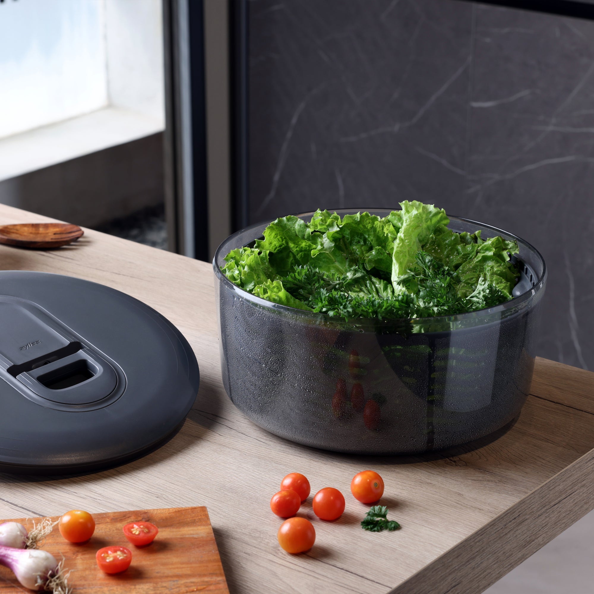 A salad spinner filled with fresh leafy greens sits on a wooden kitchen counter next to its lid, with cherry tomatoes and vegetables scattered nearby.
