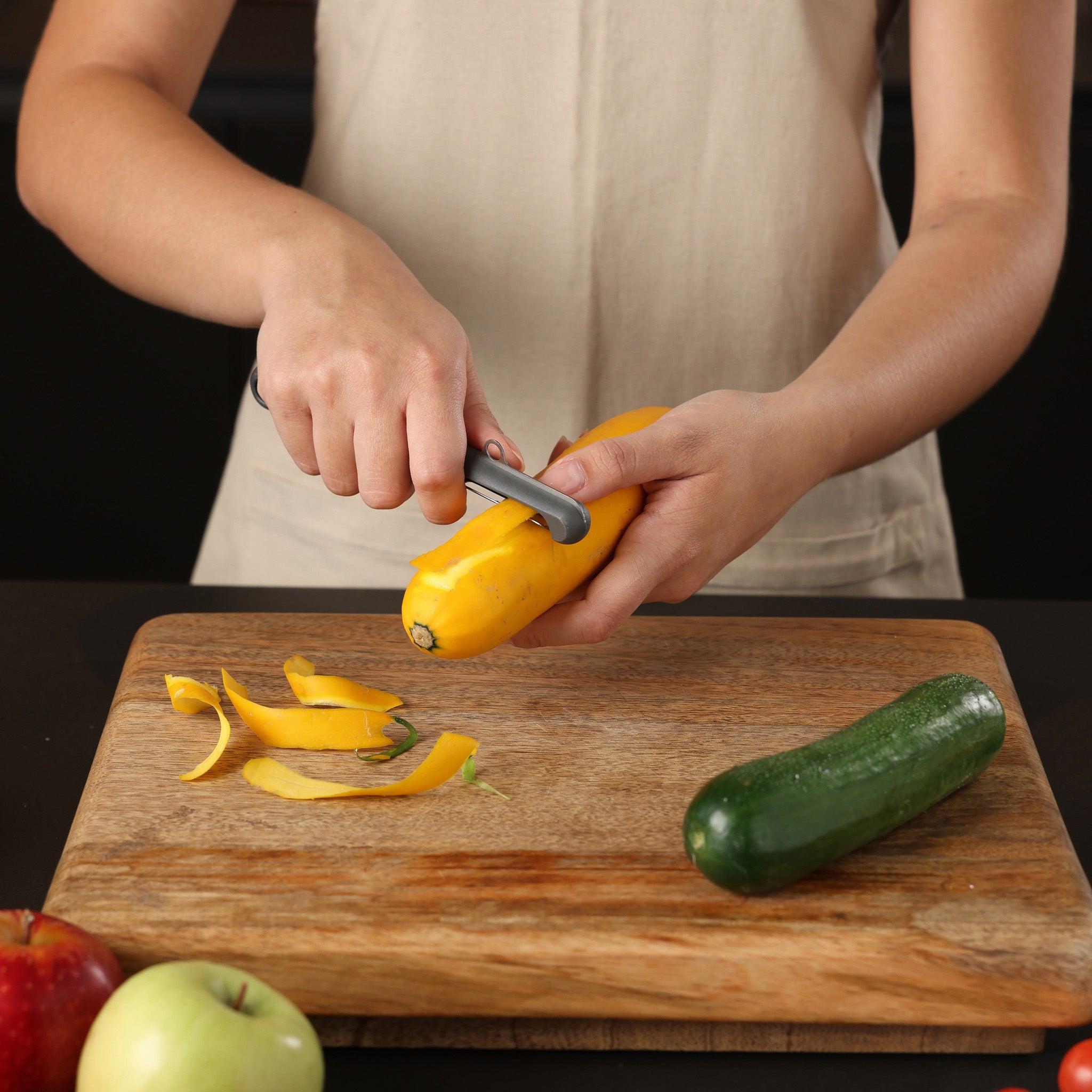 A person wearing a beige apron peels a yellow squash over a wooden cutting board. Beside the board are a green zucchini, an apple, and a peeled strip from the squash.