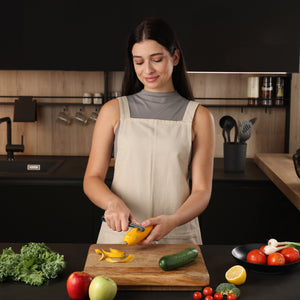 A woman in a beige apron uses the Zyliss Swivel Peeler to peel a yellow mango at her kitchen counter, which is surrounded by fresh apples, tomatoes, lemon, kale, and cucumber. 