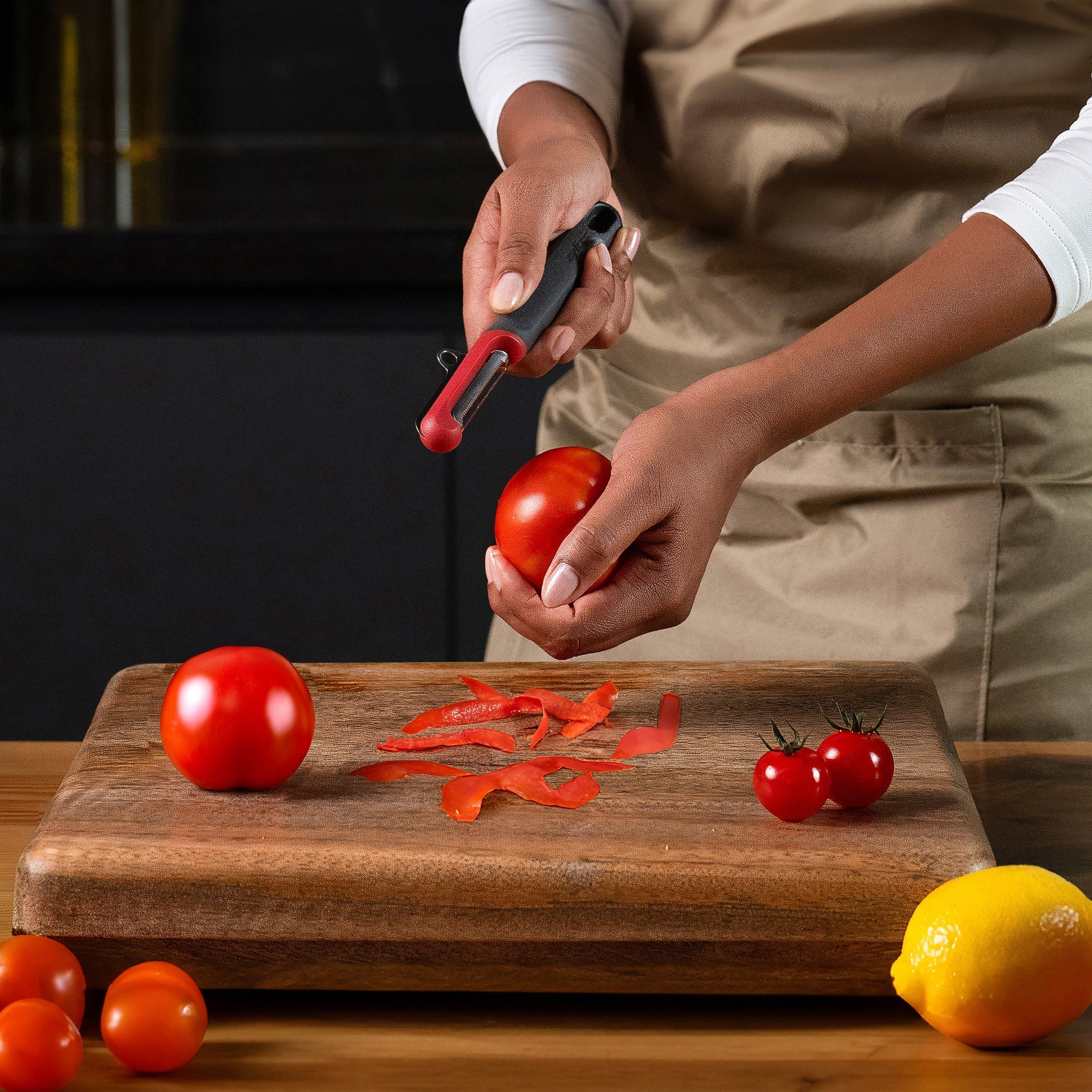 A person wearing a beige apron peels a tomato with a peeler over a wooden cutting board. The board holds tomato peels, whole tomatoes, cherry tomatoes, and a lemon.