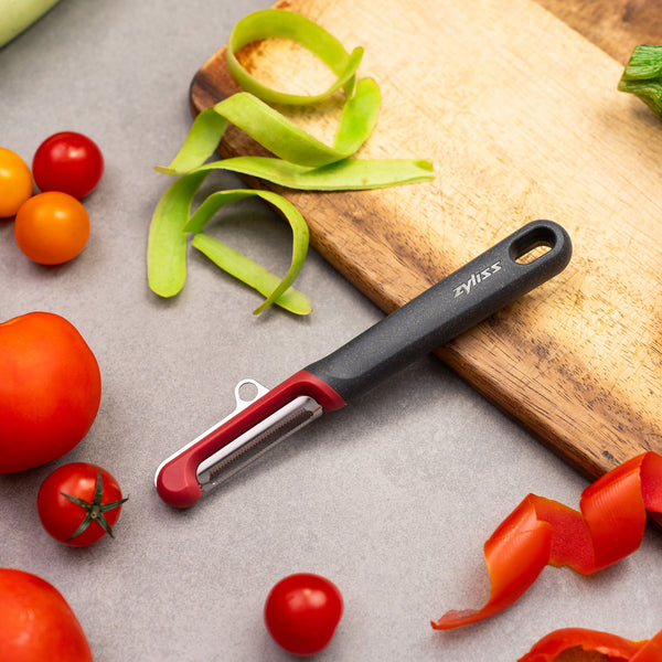The Zyliss Soft Skin Peeler, featuring a black and red handle and stainless steel blade, is displayed on a cutting board with peeled vegetable skins, tomatoes, cherry tomatoes, and a zucchini on a gray surface. 