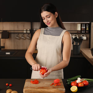 A woman in a beige apron uses the Zyliss Soft Skin Peeler with a stainless steel blade to peel a tomato in a modern kitchen. In front of her, a cutting board holds tomatoes, cucumbers, and lemon on the countertop. 