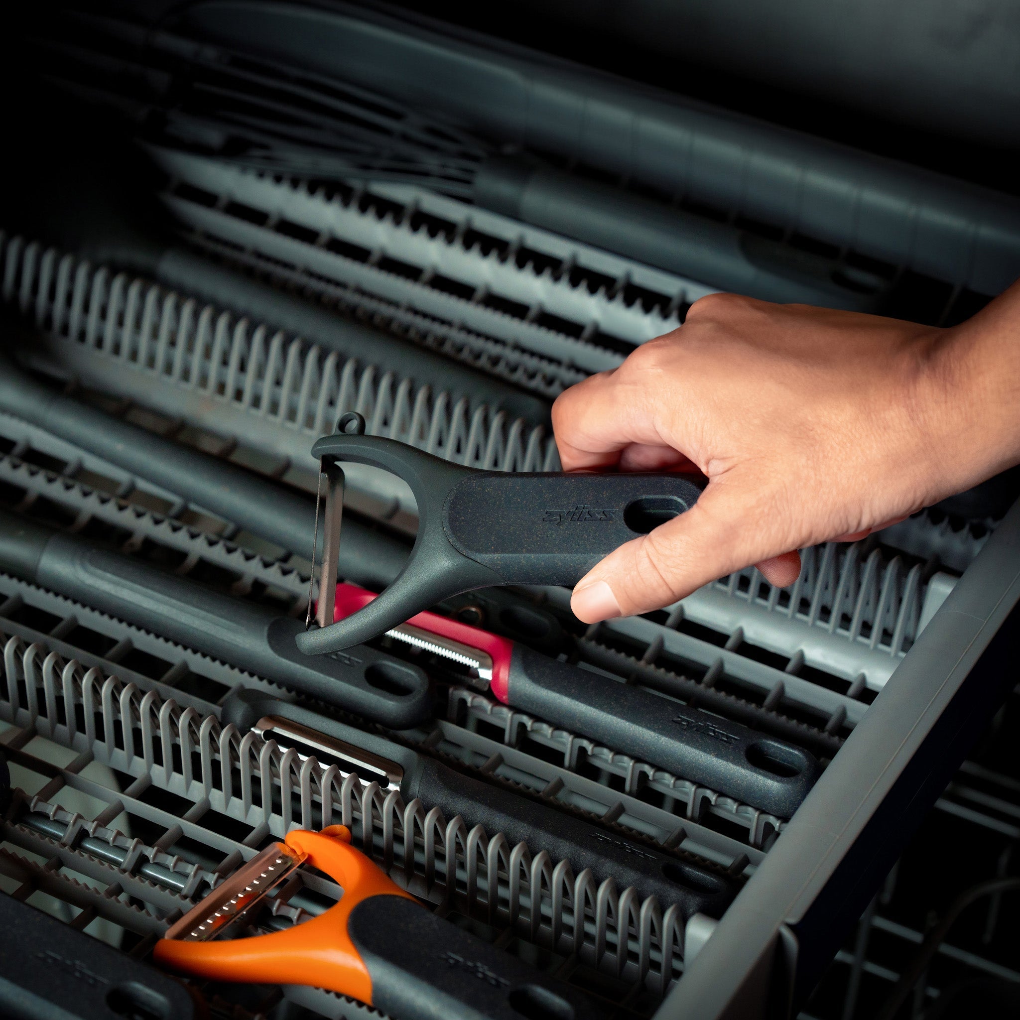 A hand is placing or removing a black vegetable peeler from a dishwasher utensil tray filled with various kitchen tools.