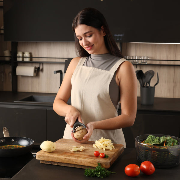A woman in a modern kitchen smiles as she peels a potato with the ergonomic Zyliss Y Peeler. She stands at a counter surrounded by a cutting board, vegetables, and fresh greens. 