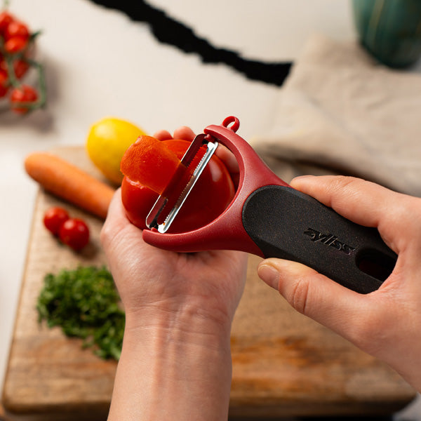 A person uses a red and black vegetable peeler to peel a tomato over a wooden cutting board with herbs, a lemon, a carrot, and tomatoes in the background.