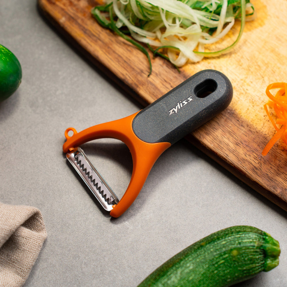 The Zyliss Julienne Y Peeler features an orange handle, gray ergonomic grip, and a horizontal stainless steel blade, displayed on a plain white background. 