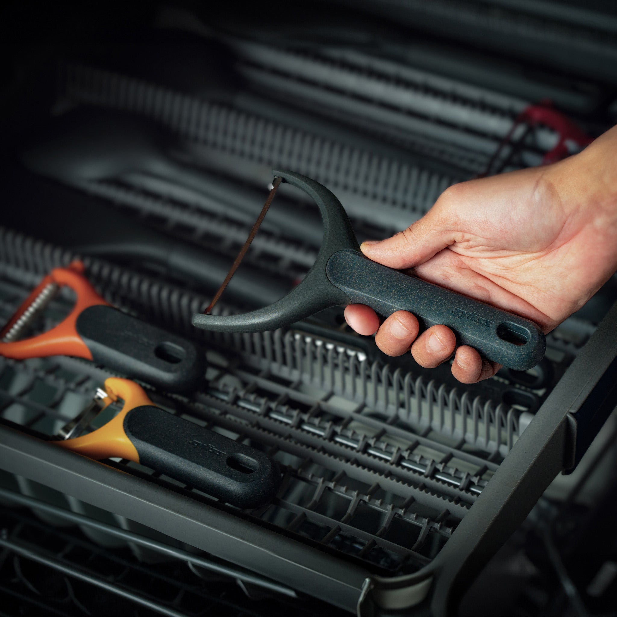 A hand places a black-handled vegetable peeler into the top rack of a dishwasher, where two other peelers with different colored handles are also visible.