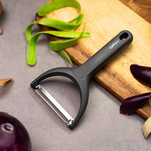 A Zyliss Wide Peeler with an ergonomic handle sits on a gray countertop next to a wooden cutting board, surrounded by green and purple vegetable skins. 