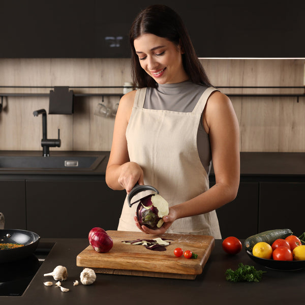 A woman in a modern kitchen smiles as she peels an eggplant with the ergonomic Zyliss Wide Peeler. Fresh tomatoes, zucchini, mushrooms, and parsley sit on the counter beside her. 