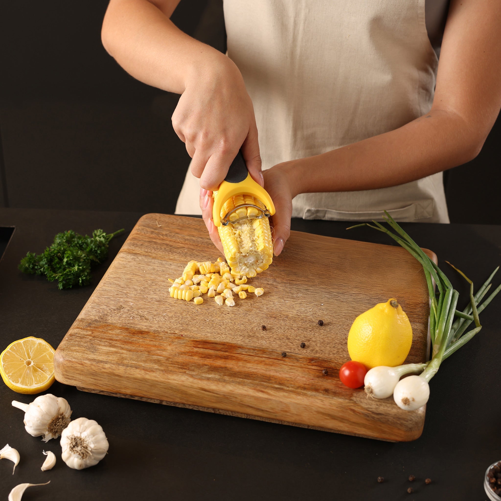 Wearing an apron, a person uses the Zyliss Corn Stripper with an ergonomic handle to remove kernels from a cob over a wooden cutting board surrounded by lemon, garlic, green onions, tomatoes, and fresh herbs.