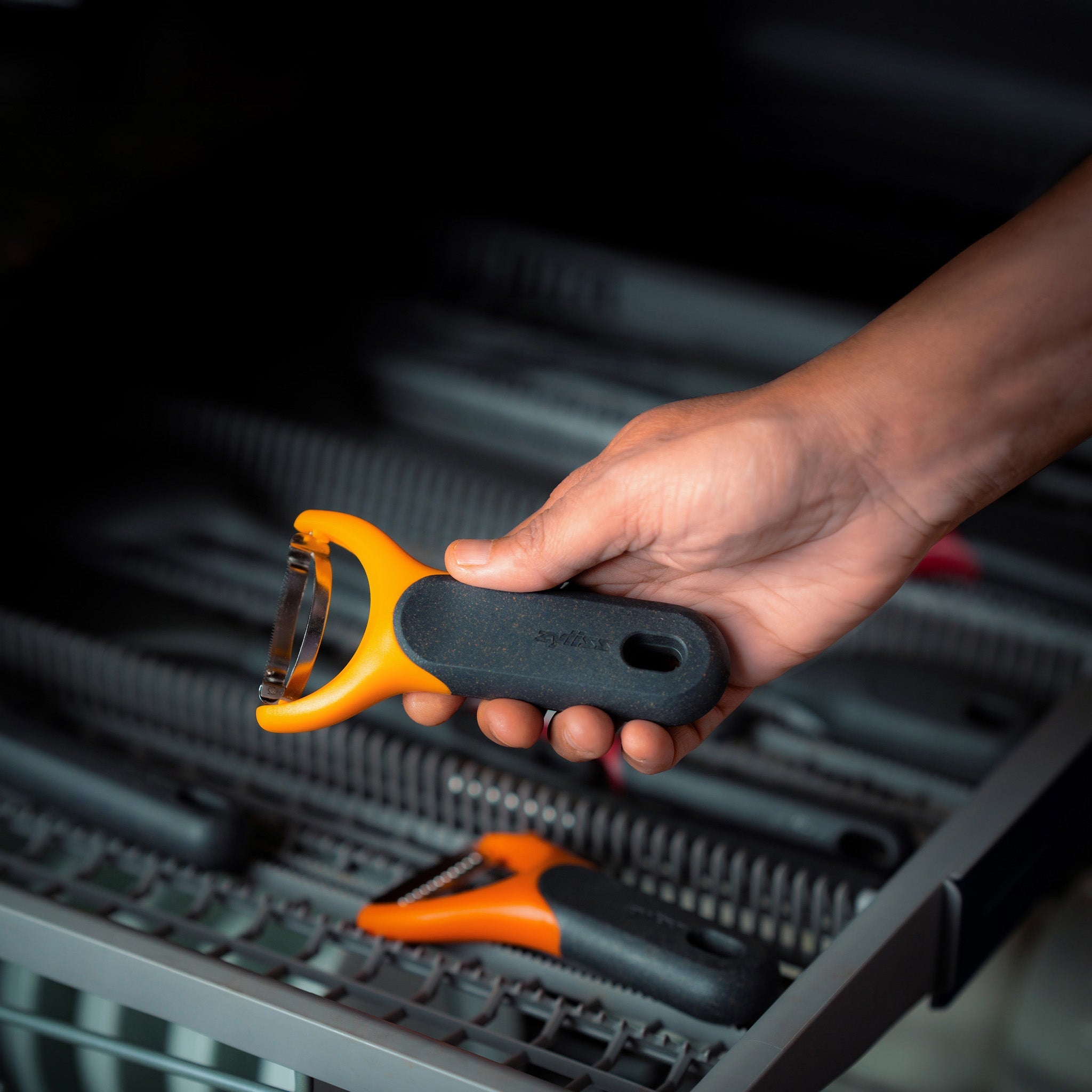A hand holding an orange and black kitchen tool above a dishwasher rack, with similar tools lying inside the dishwasher.