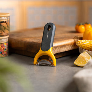 A yellow and black Zyliss Corn Stripper with an ergonomic handle stands upright on a kitchen counter near a lemon wedge, corn, and a wooden cutting board, with jars and vegetables in the background. 