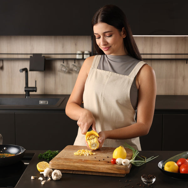 A woman in a beige apron stands in a modern kitchen, squeezing a lemon over veggies and spices next to the Zyliss Corn Stripper, an ergonomic tool for easy kernel removal on the wooden cutting board. 
