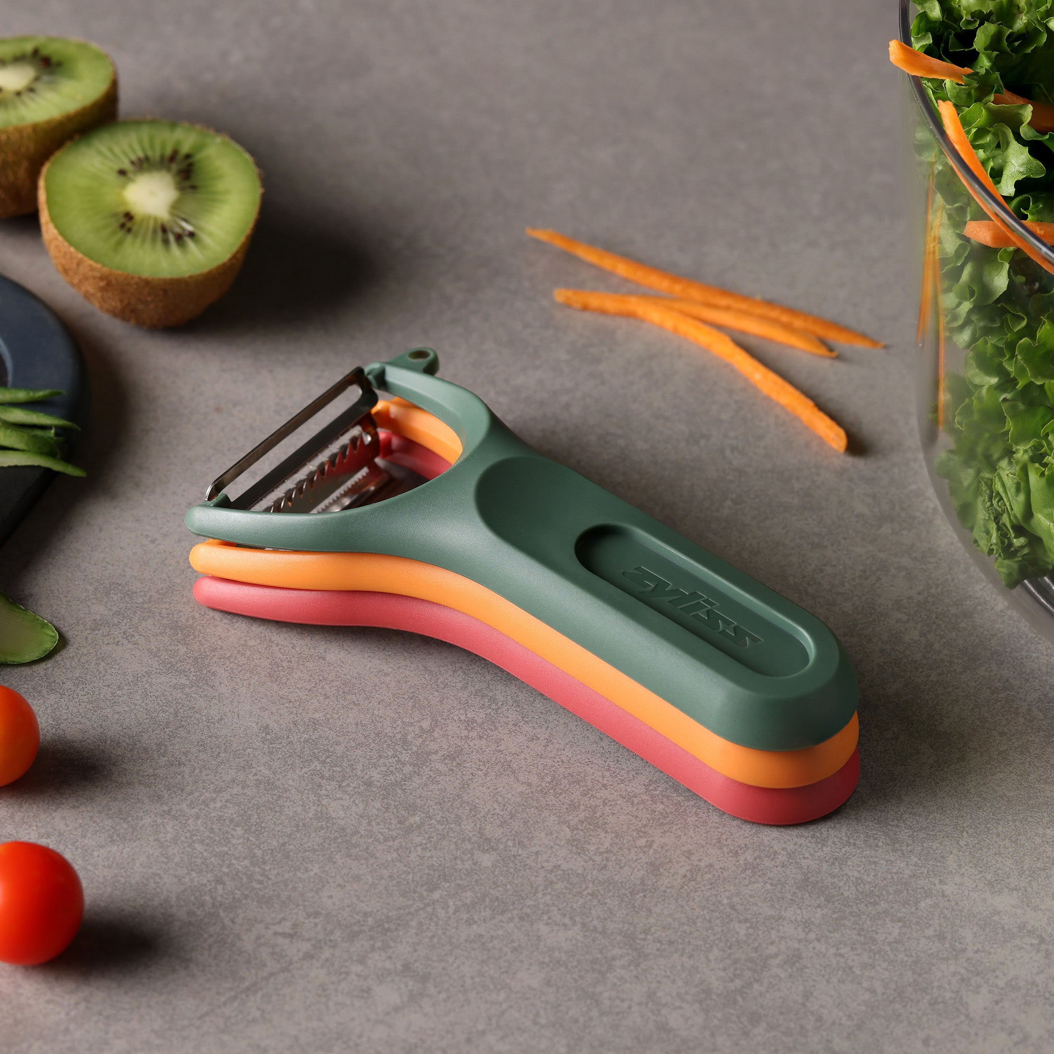 The Zyliss 3 Piece Y Peeler Set in green, orange, and pink rests on a gray countertop beside sliced kiwi, carrot sticks, leafy greens, cherry tomatoes, and cucumber slices.