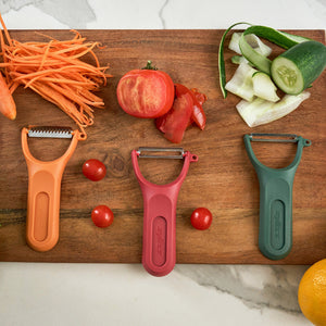A Zyliss 3 Piece Y Peeler Set in orange, red, and green is displayed on a wooden board with peeled carrots, tomatoes, cucumber, lemon, vegetable peels, and cherry tomatoes on a marble counter.