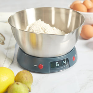 A metal bowl with flour sits on a Zyliss Digital Kitchen Scale showing 614g with tare for accuracy; lemons, figs, and eggs are nearby on the white countertop. 