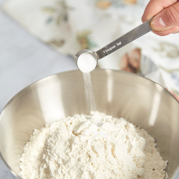 A hand sprinkles a teaspoon of white powder into a metal bowl of flour using Zyliss Stainless Steel Measuring Spoons—an essential kitchen tool. A blurred floral cloth appears in the background. 