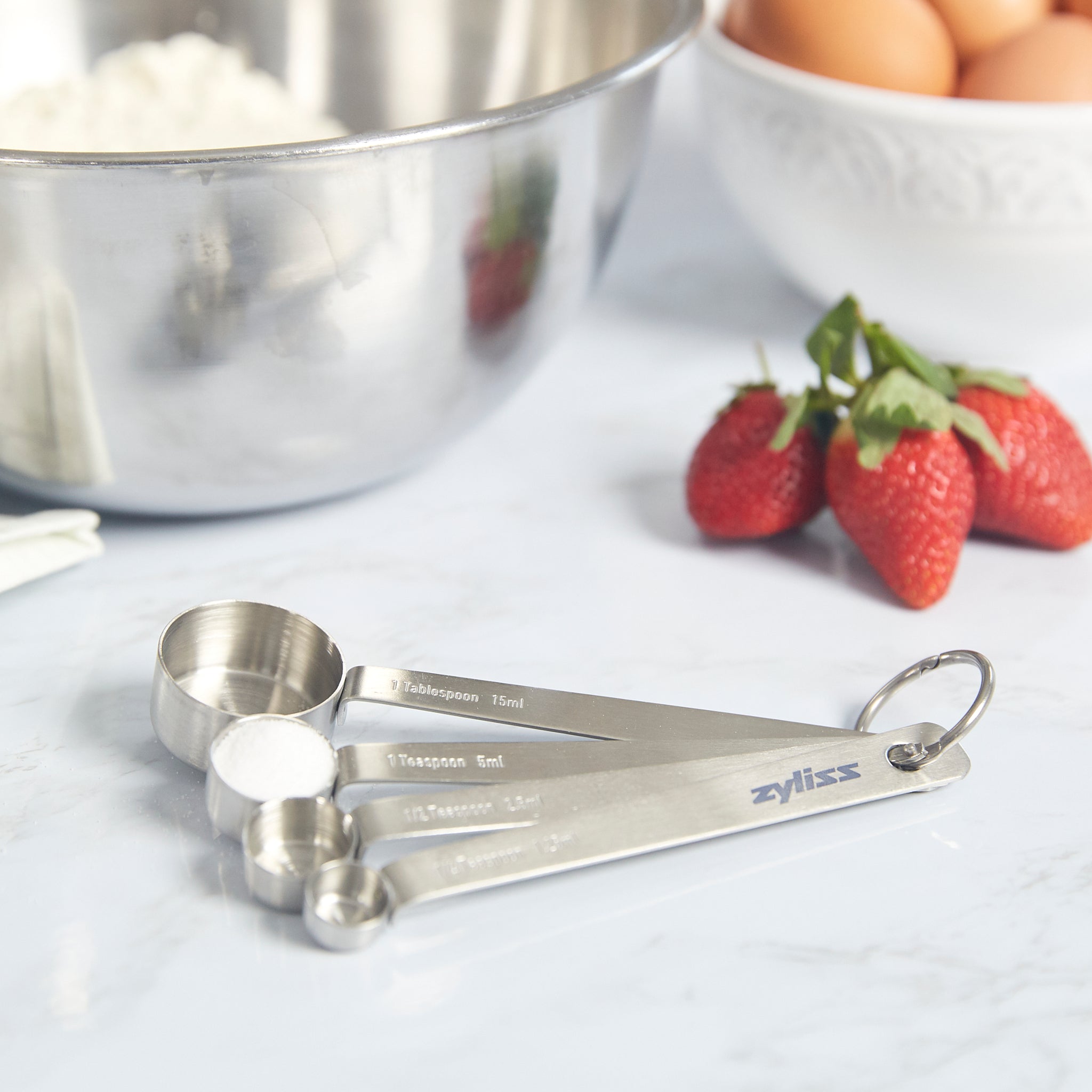 The Zyliss Stainless Steel Measuring Spoons—a kitchen essential—sit on the countertop next to fresh strawberries, eggs, and a stainless mixing bowl filled with flour, perfectly ready for your next baking or cooking task.