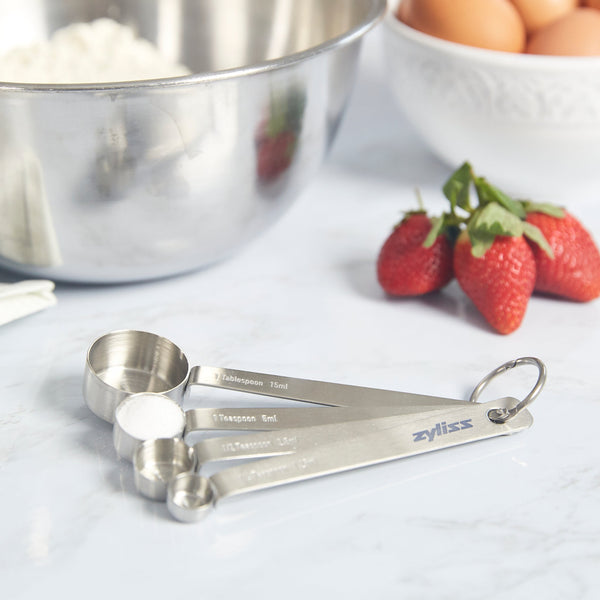 The Zyliss Stainless Steel Measuring Spoons—a kitchen essential—sit on the countertop next to fresh strawberries, eggs, and a stainless mixing bowl filled with flour, perfectly ready for your next baking or cooking task. 