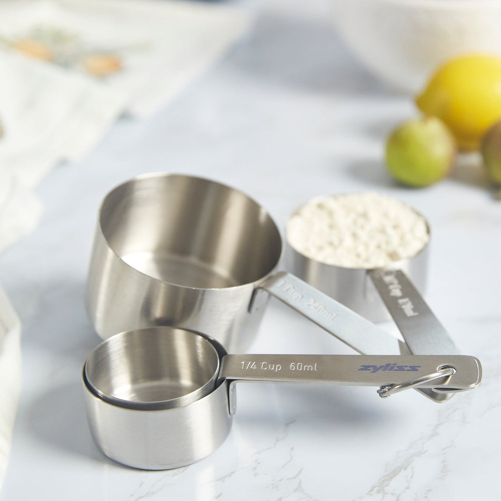 The Zyliss Stainless Steel Measuring Cups set includes four stacked, fanned-out cups, each labeled in cups and milliliters for precise ingredient measurement. 