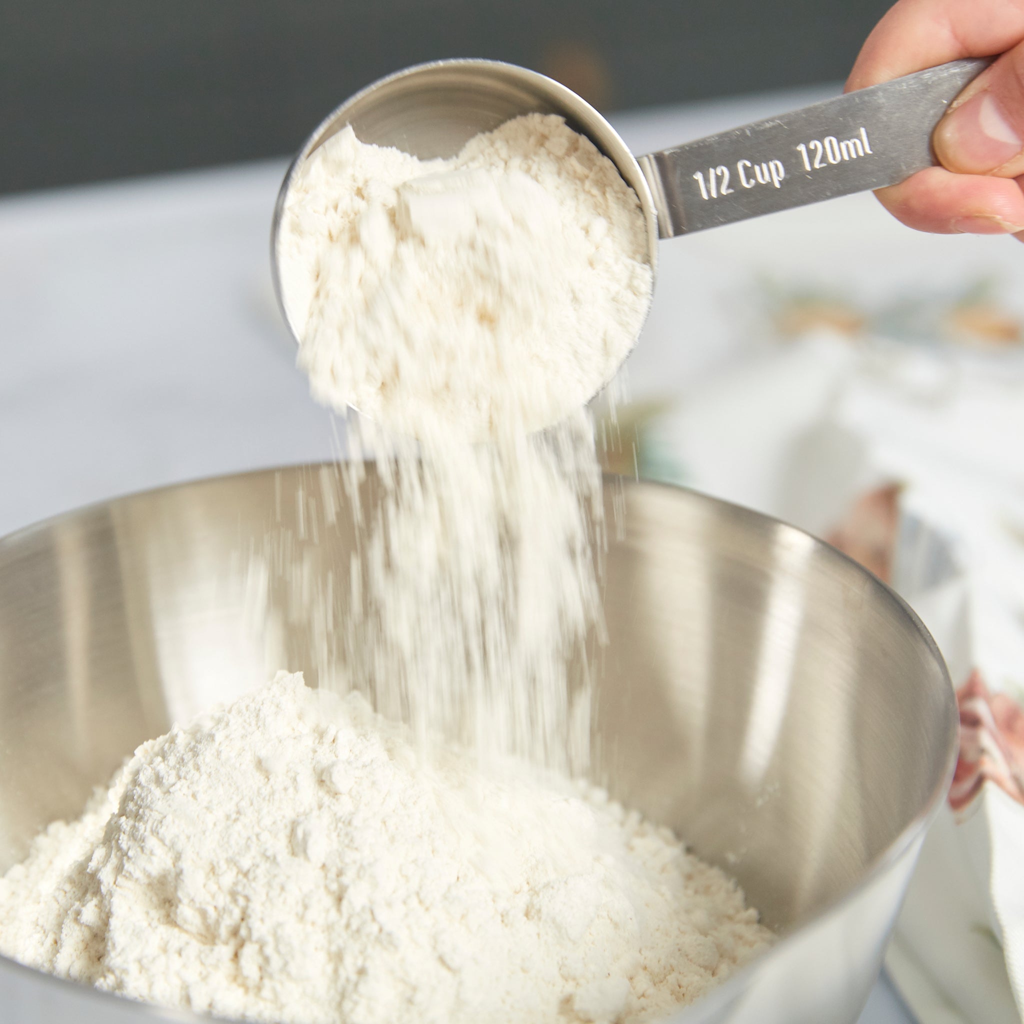A Zyliss Stainless Steel Measuring Cup labeled 1/2 Cup 120 ml pours flour into a mixing bowl, demonstrating precise ingredient measurement. A softly blurred background accentuates this essential kitchen tool in use.